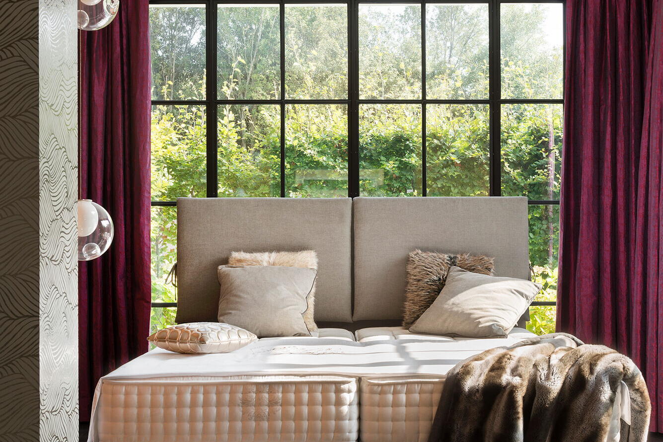 Boxspringbett mit grauem Kopfteil vor einer Fensterfront