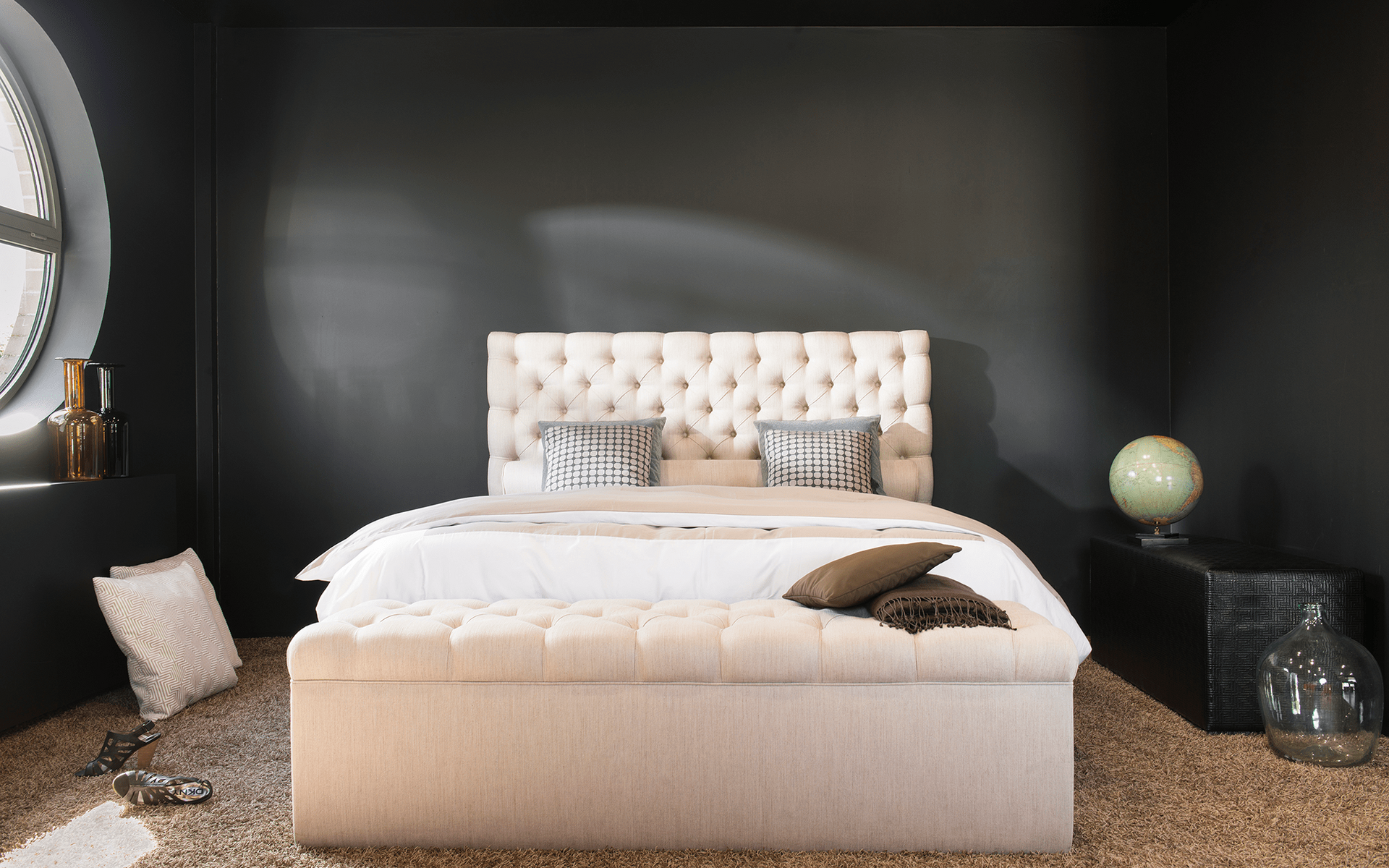 Cremefarbenes Boxspringbett mit geknöpftem Kopfteil vor dunkler Wand im Showroom von Trend Base